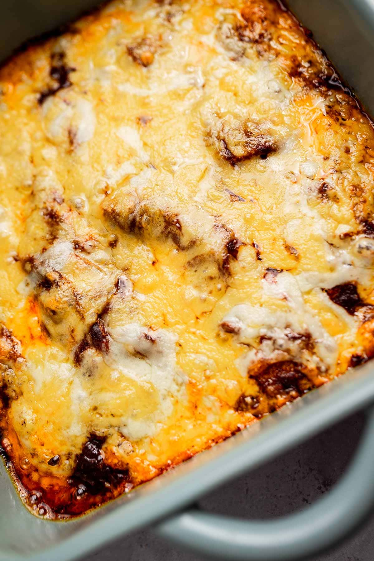 A close-up of a cheese enchiladas casserole showcases the oozing melted cheese and browned edges in a rectangular baking dish.