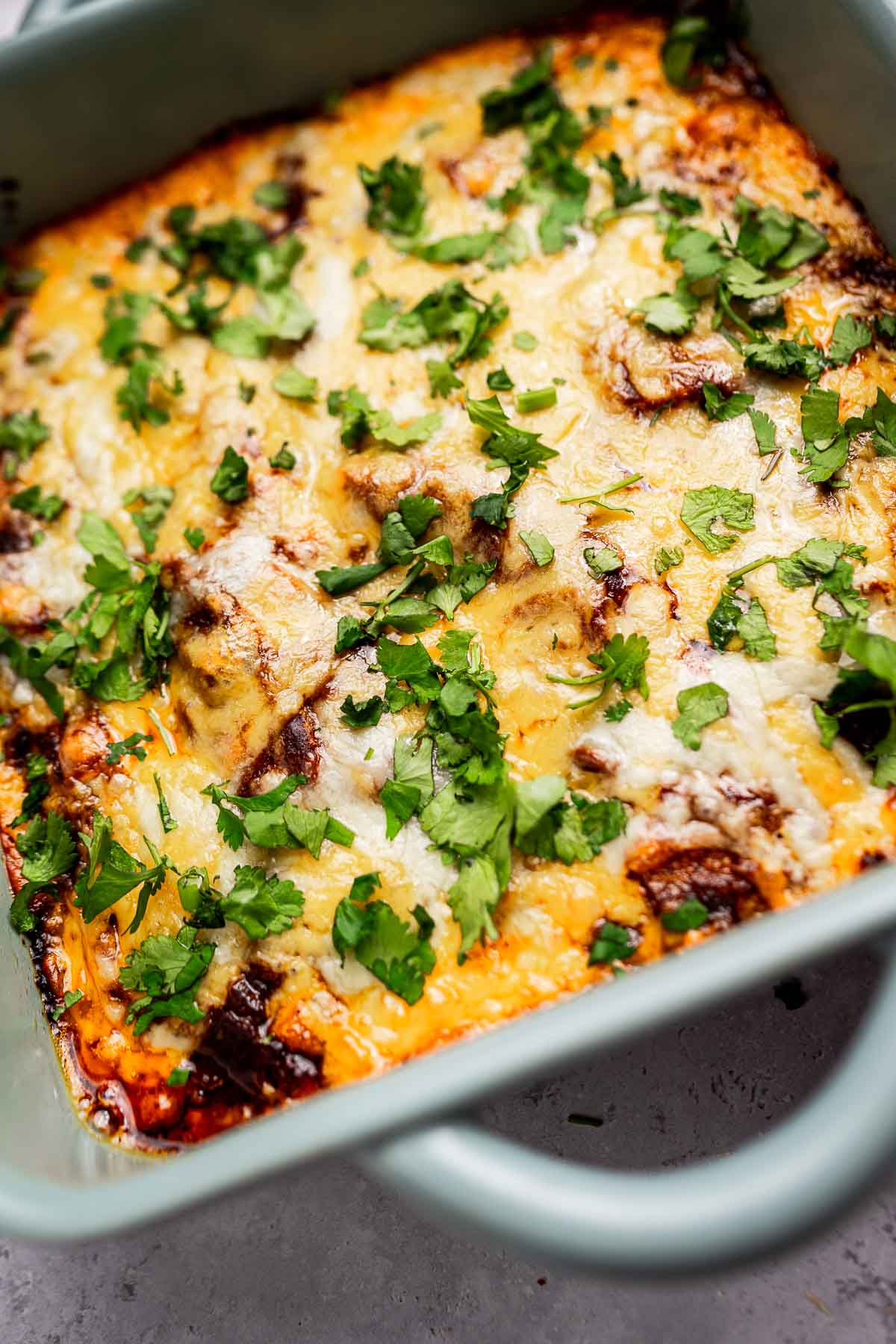 A baked cheese enchiladas casserole in a green dish, topped with melted cheese and sprinkled with chopped cilantro.