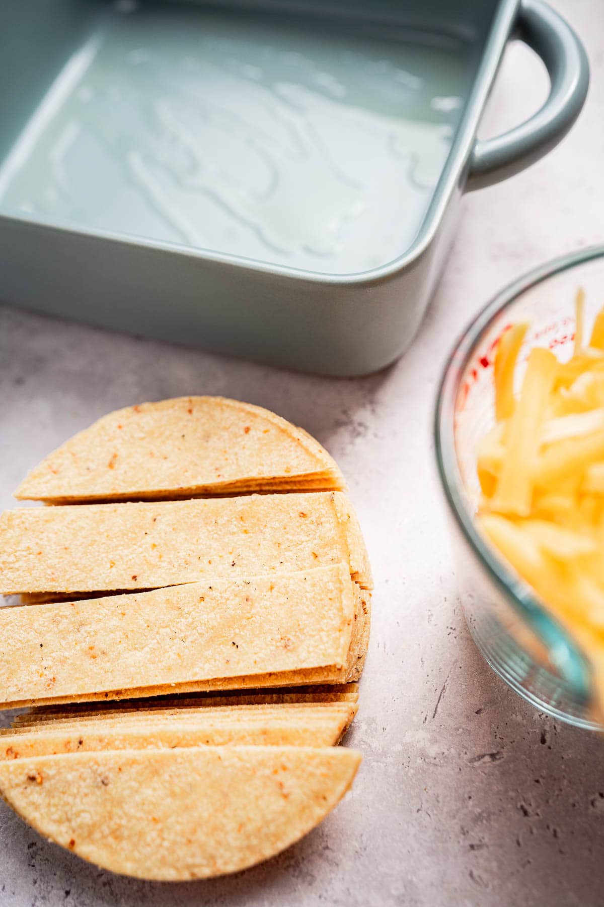 Stack of sliced tortillas beside a green baking dish and a bowl of shredded cheese on a countertop, ready to be transformed into a delicious cheese enchiladas casserole.