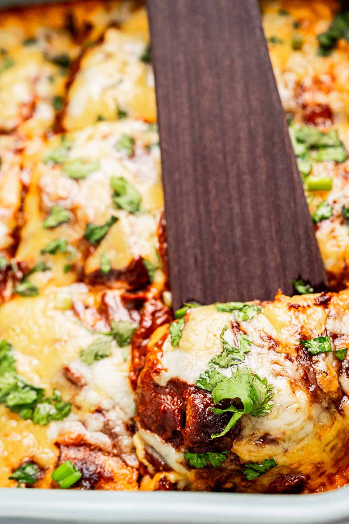 A close-up of a cheese enchiladas casserole with melted cheese, garnished with herbs. A spatula lifts a portion, revealing the savory layers of filling underneath.