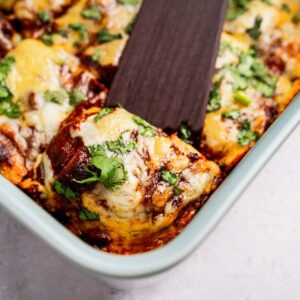 A spatula lifts a cheesy, baked enchilada topped with cilantro from a baking dish.