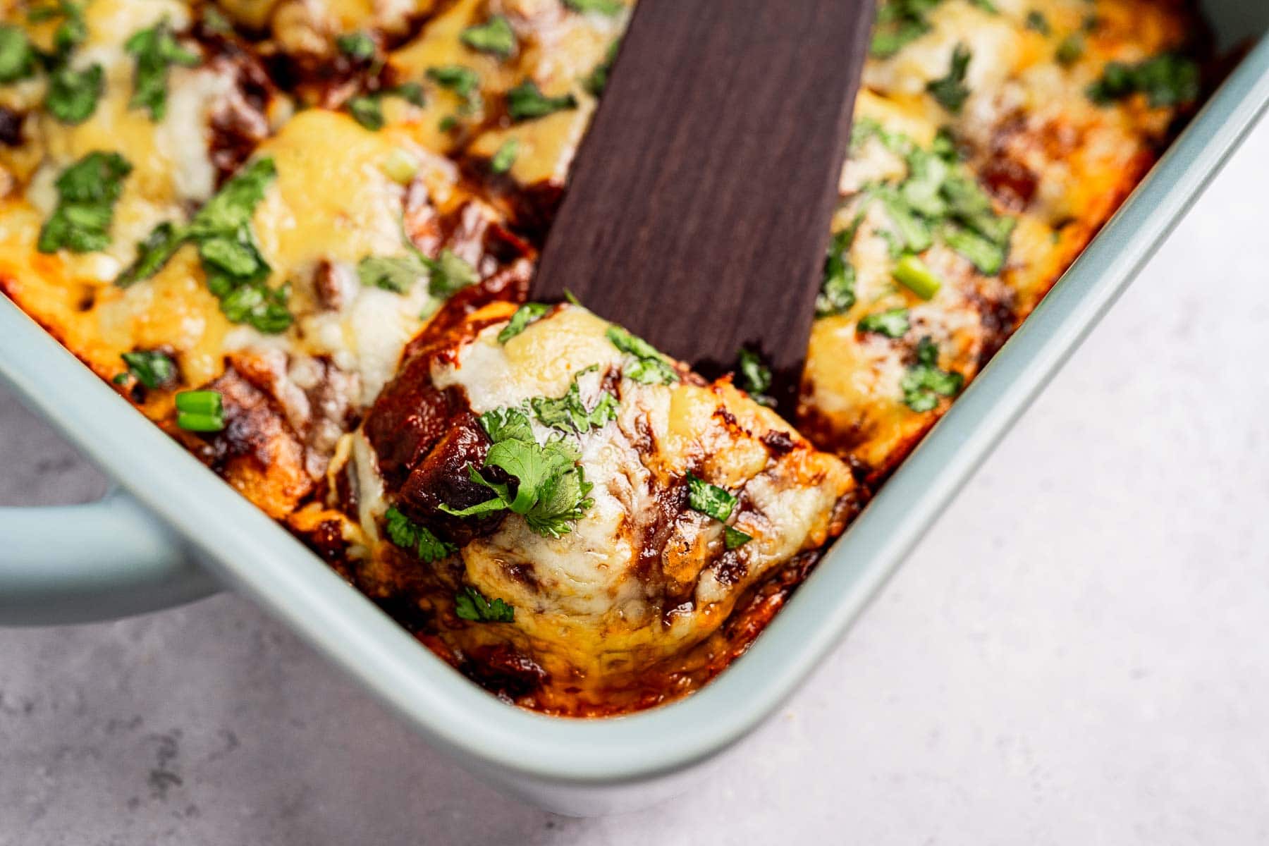 A close-up of a cheese enchiladas casserole in a dish, topped with melted cheese and chopped cilantro. A spatula is lifting a portion.