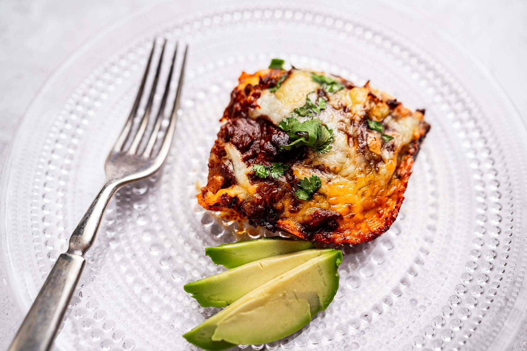 A slice of lasagna, reminiscent of a cheese enchiladas casserole, is garnished with cilantro on a clear glass plate, accompanied by avocado slices. A fork rests nearby.