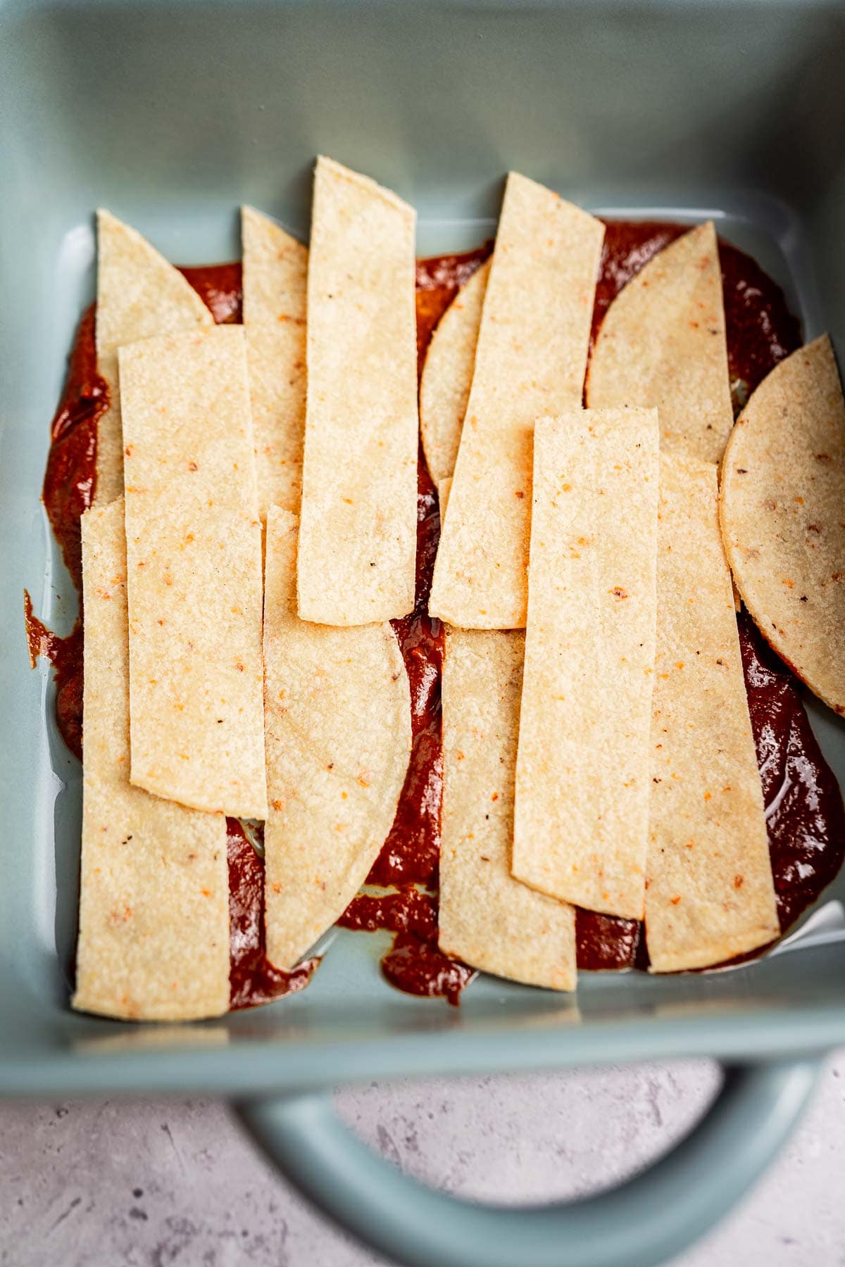 Strips of tortilla lay harmoniously over a layer of zesty red sauce in a rectangular baking dish, creating the perfect base for this cheesy enchiladas casserole.
