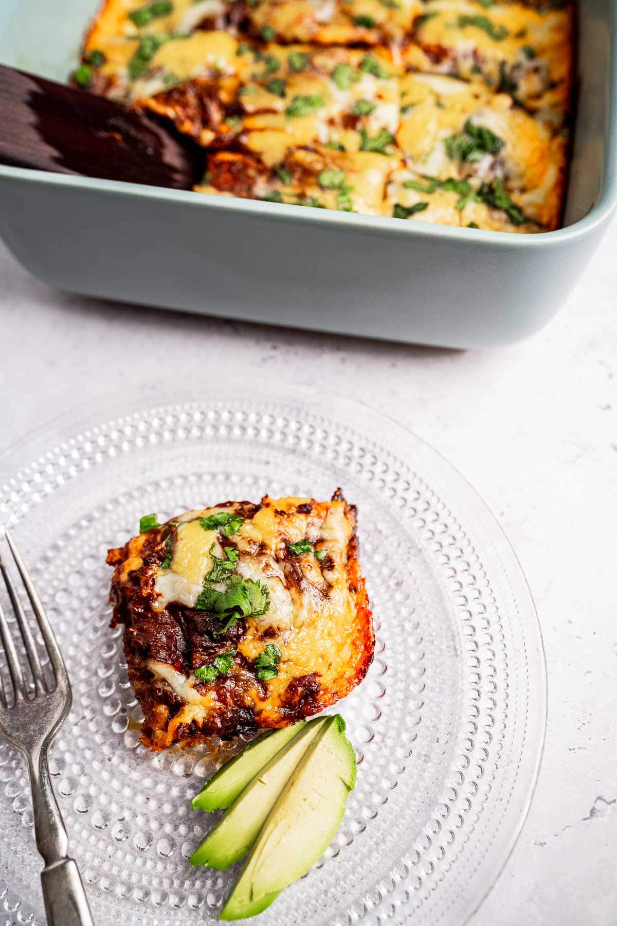 A slice of cheesy enchilada casserole rests on a clear plate, garnished with creamy avocado slices, sitting beside a baking dish brimming with more of the savory delight.
