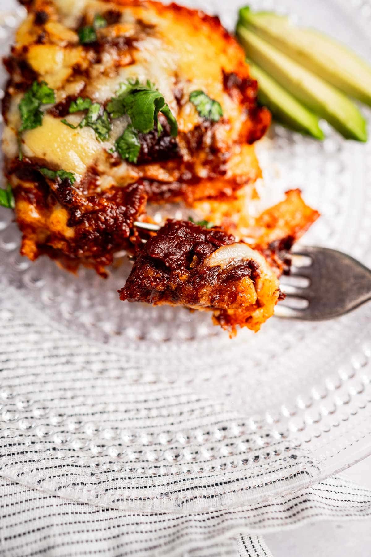 A serving of cheese enchiladas casserole topped with melted cheese and cilantro on a glass plate. A fork lifts a portion, with sliced avocado in the background.