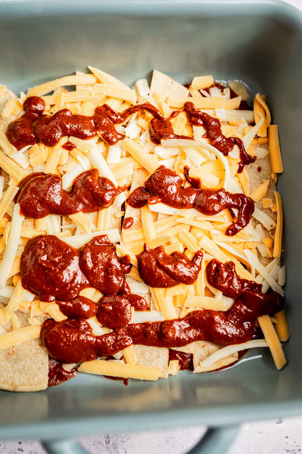 A baking dish layered like cheese enchiladas, featuring tortilla chips, melted cheese, and dollops of sauce.