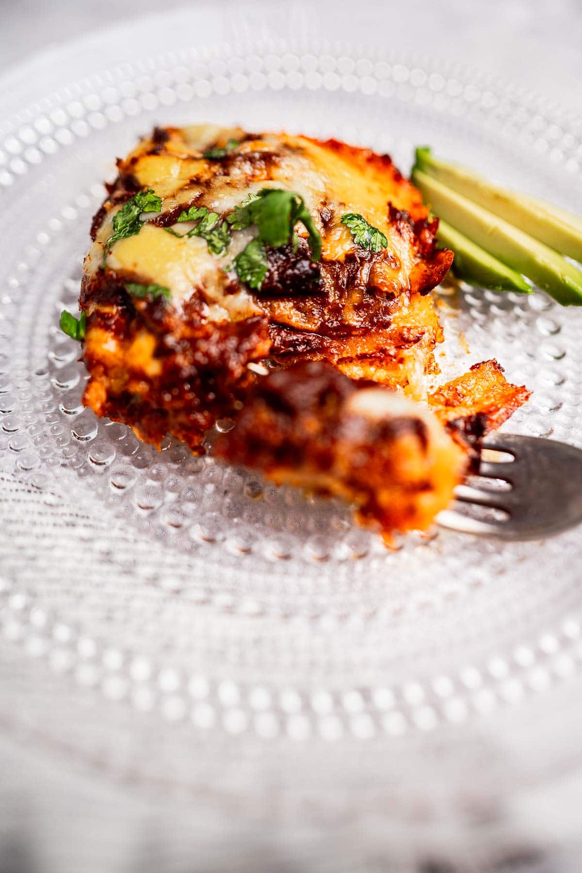 Cheesy, baked chicken breast on a glass plate, reminiscent of a savory cheese enchiladas casserole, topped with herbs and served with sliced avocado. A fork is placed beside the chicken.