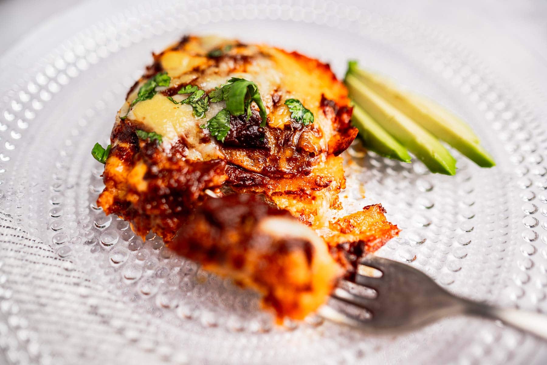 Close-up of a portion of lasagna topped with melted cheese and herbs on a clear plate, reminiscent of a savory cheese enchiladas casserole. Accompanied by slices of avocado, with a fork invitingly placed in front.