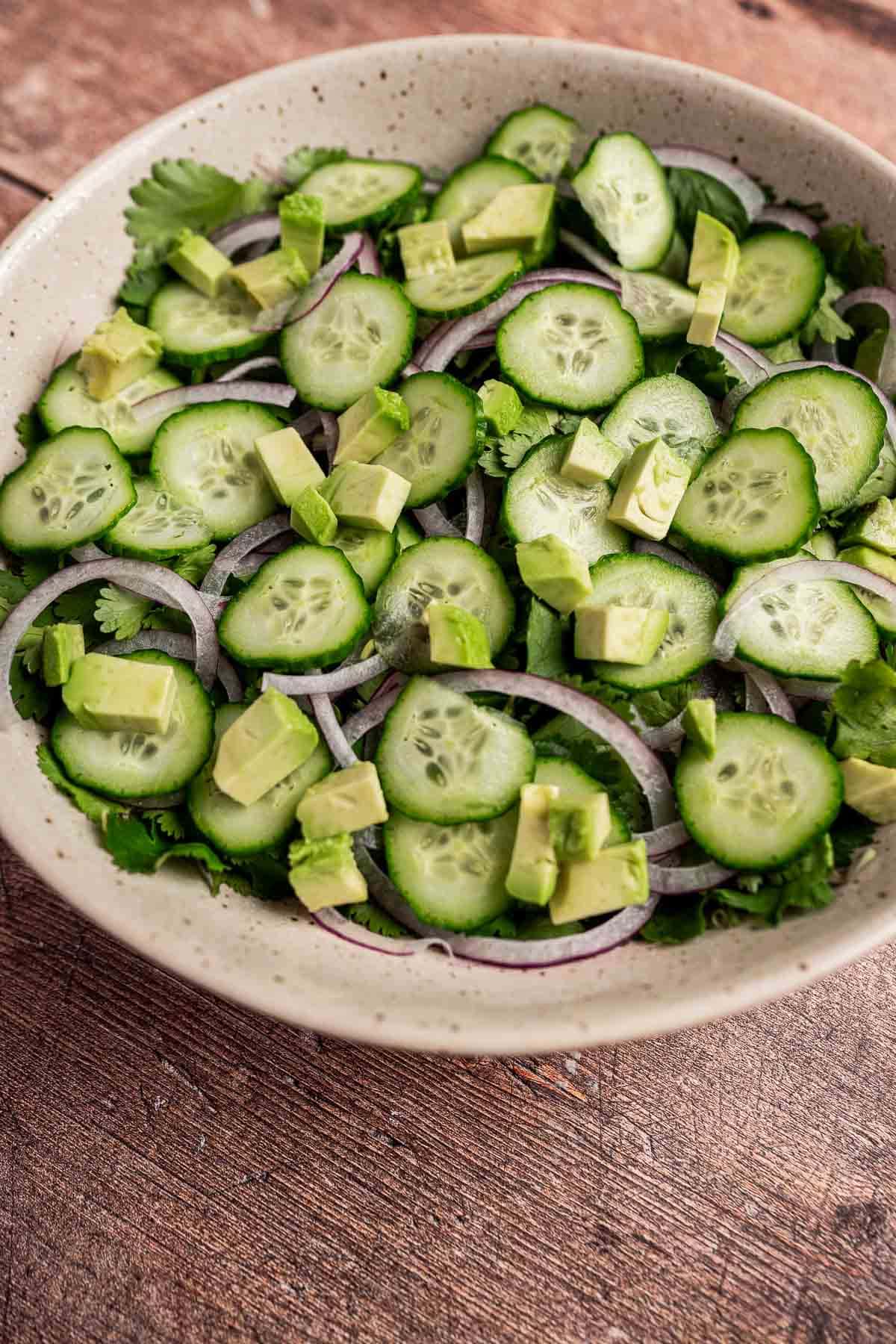 A bowl of cilantro salad delights with avocado chunks, crisp cucumber, and red onion slices resting on a wooden surface.