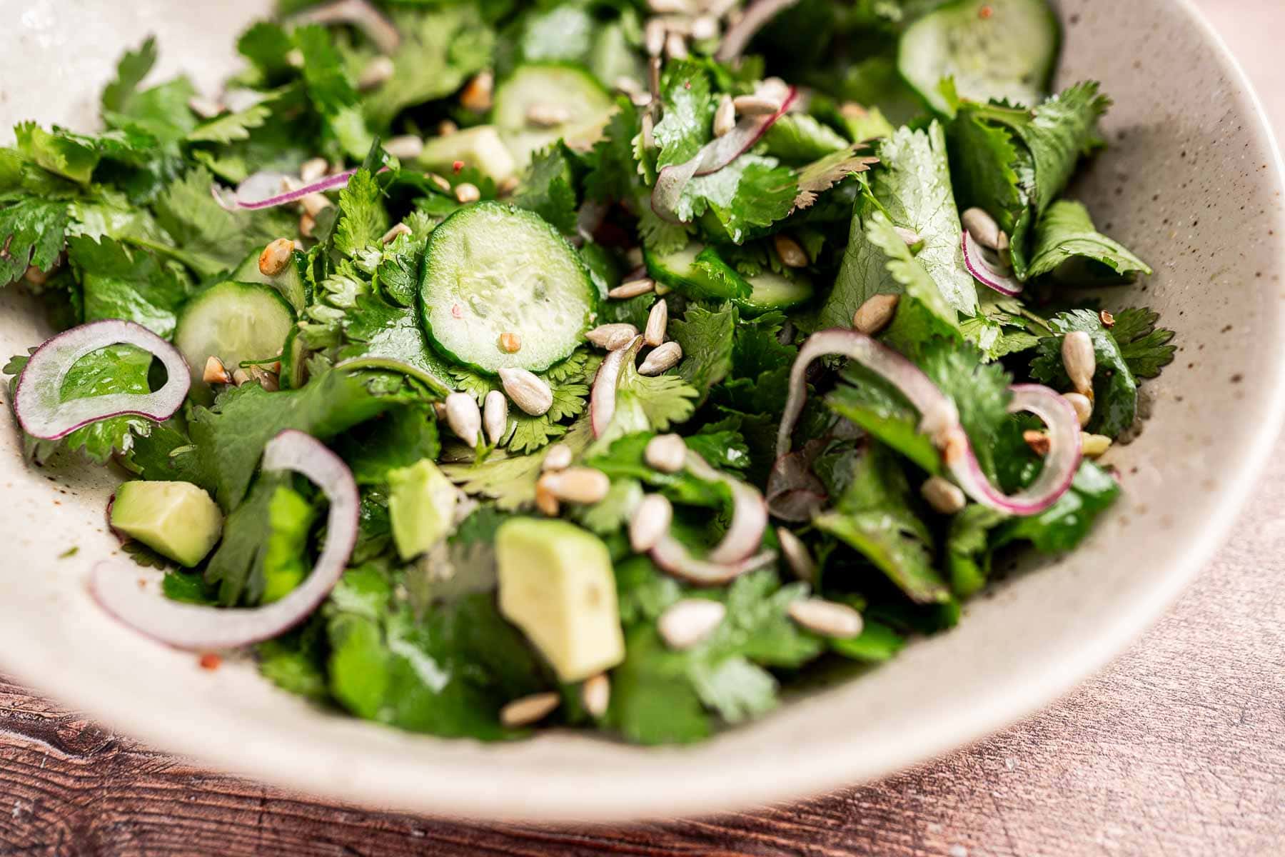 A cilantro salad featuring cucumber slices, red onion, avocado pieces, and sunflower seeds is elegantly arranged in a speckled bowl on a wooden surface.