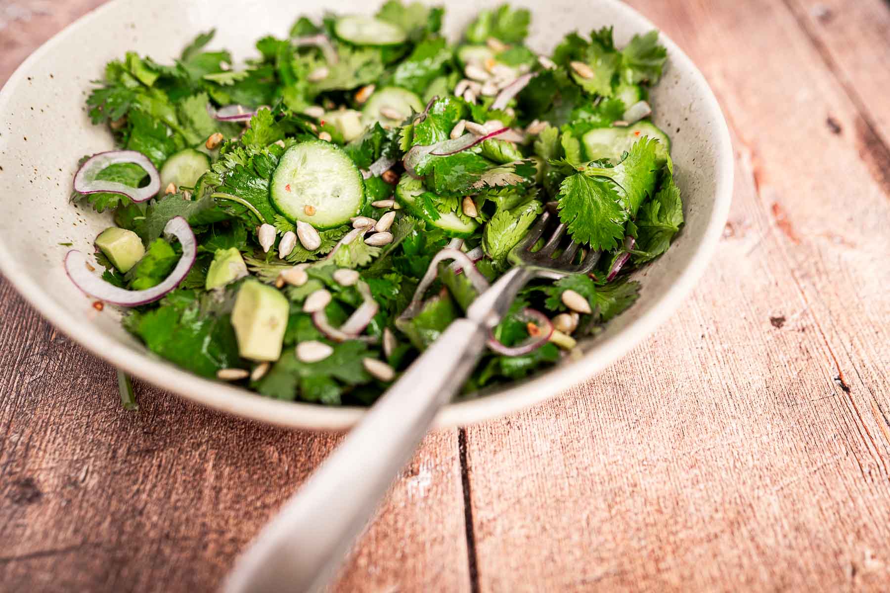 A cilantro salad with cucumber slices, red onion, avocado, and sunflower seeds sits invitingly on a wooden surface, with a fork nestled inside the bowl.