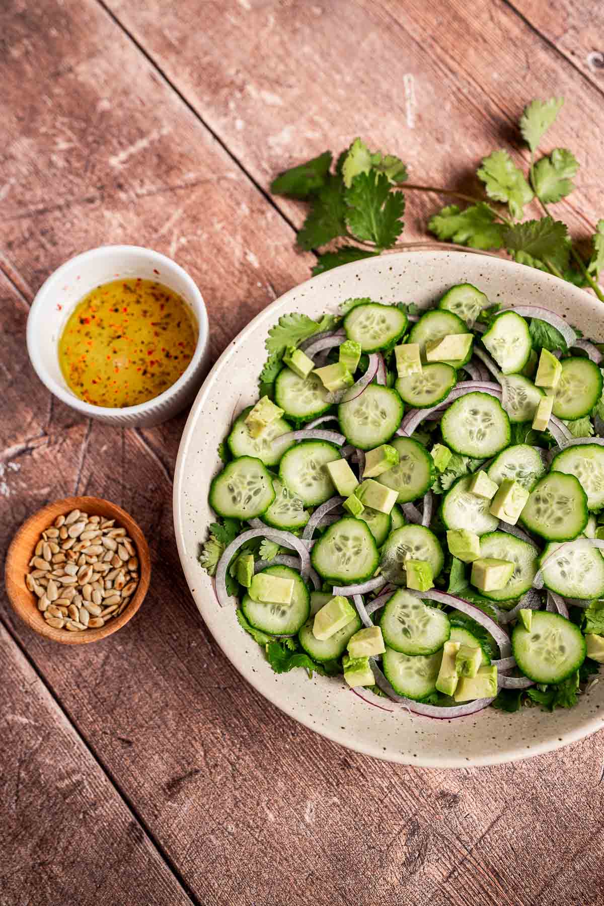 A bowl of cilantro salad featuring cucumber, avocado, and red onion sits on a wooden table. Nearby are a small dish of dressing and a bowl of sunflower seeds for added flavor.