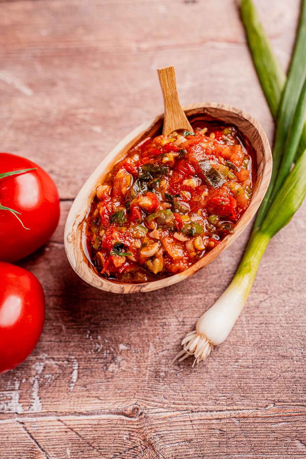 A wooden bowl of salsa hogao with a spoon, surrounded by two tomatoes and green onions on a wooden surface.