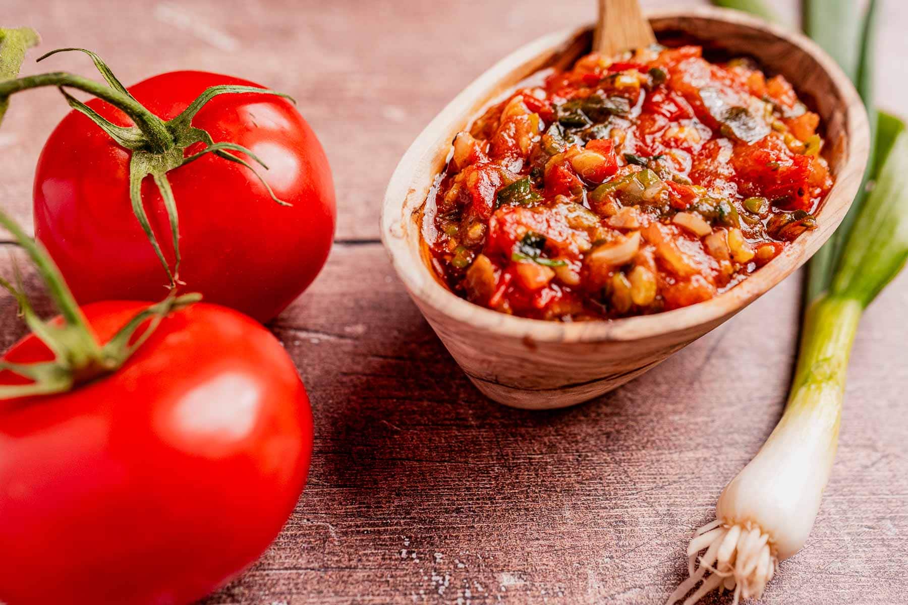 A wooden bowl of hogao with a wooden spoon rests beside two ripe tomatoes and a green onion on a rustic wooden surface.