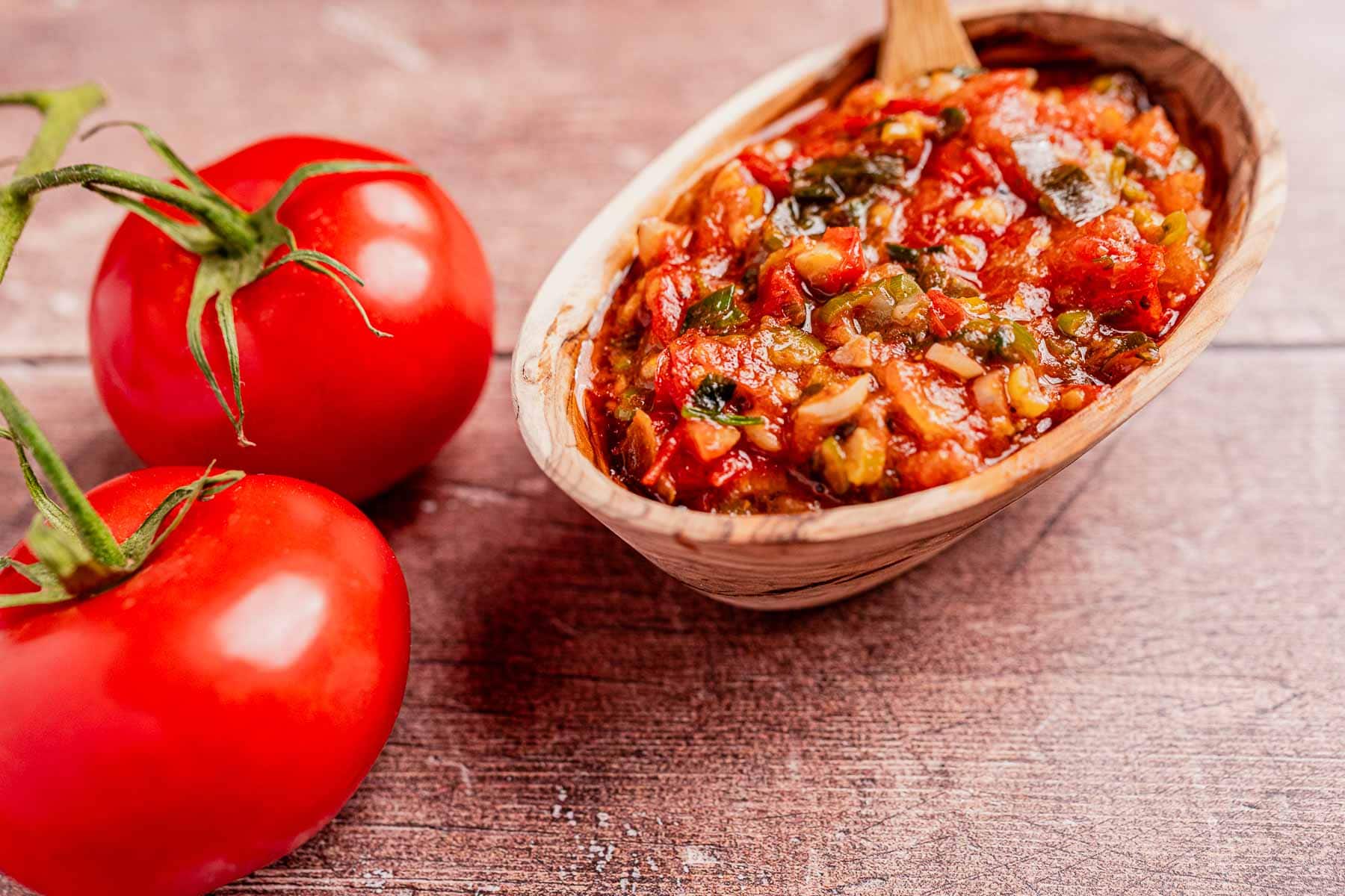 A wooden bowl filled with hogao, a savory tomato-based salsa, sits next to two ripe tomatoes on a rustic wooden surface.