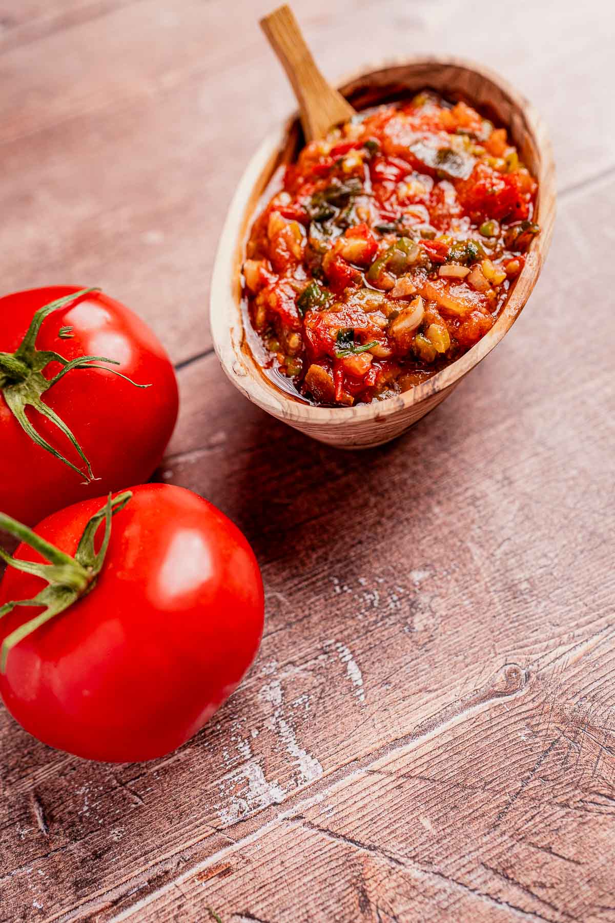 Wooden bowl with chunky salsa, reminiscent of traditional hogao, and a wooden spoon beside two vibrant red tomatoes on a rustic wooden surface.