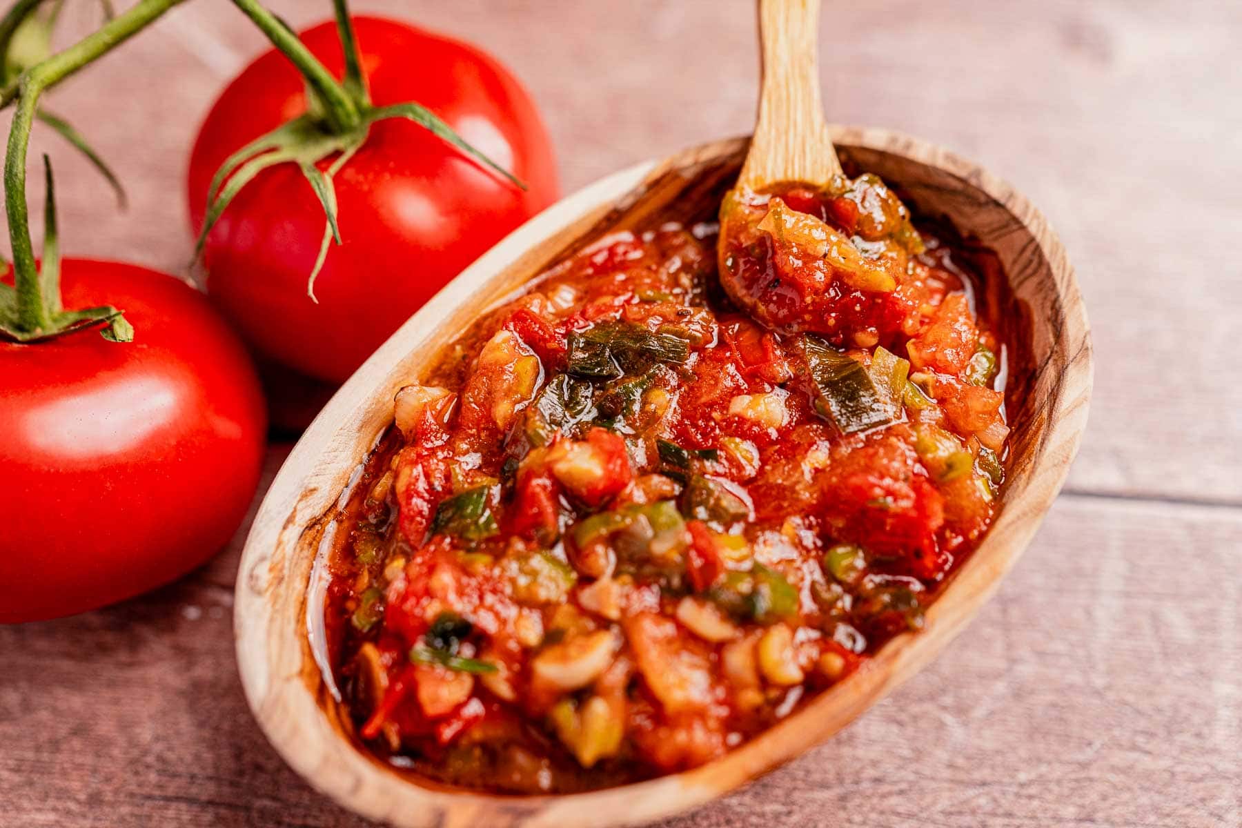 A wooden bowl brimming with chunky tomato salsa, reminiscent of traditional hogao, sits invitingly with a spoon beside two fresh vine-ripened tomatoes.