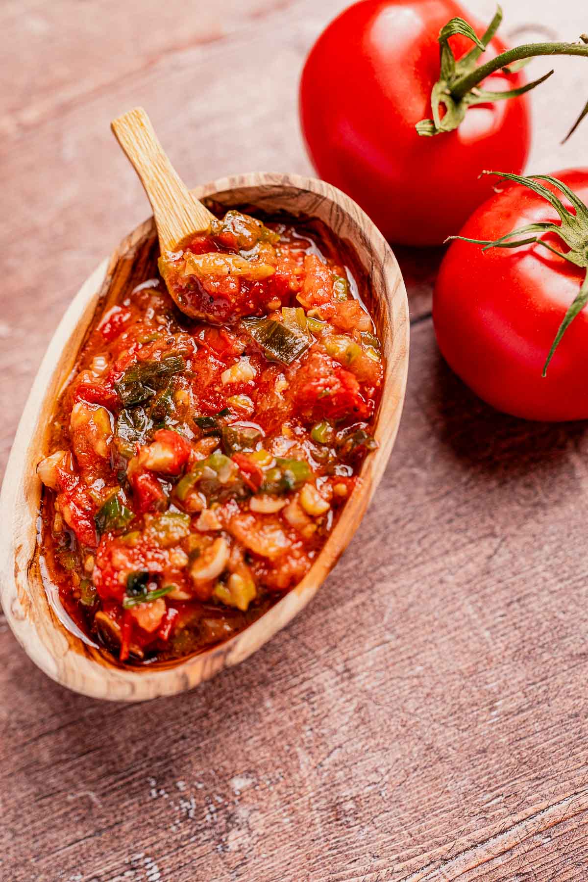 A wooden bowl filled with chunky salsa, reminiscent of traditional hogao, contains tomatoes and green peppers, complemented by a small wooden spoon. Two fresh red tomatoes sit elegantly beside the bowl on a rustic wooden surface.