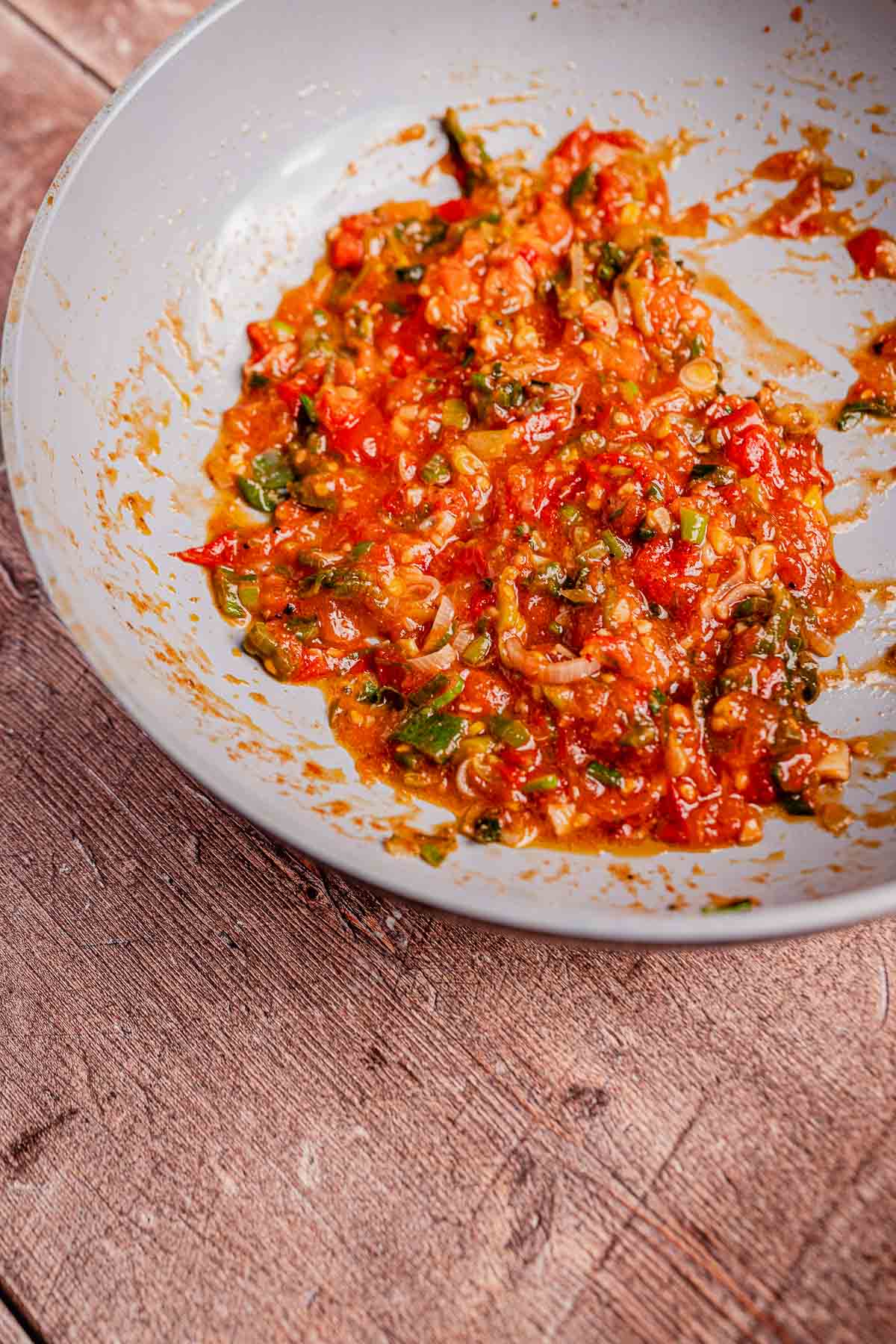 A white ceramic bowl holds a chunky, vibrant tomato-based salsa with red and green hues resembling traditional hogao, resting elegantly on a wooden surface.
