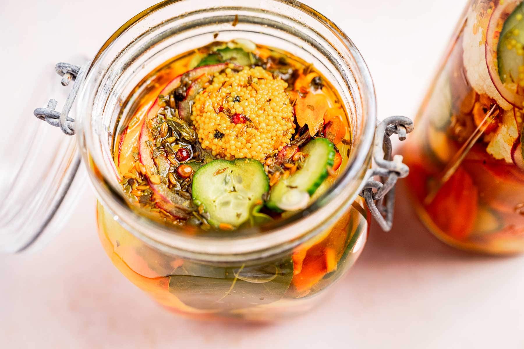 An open glass jar teems with vibrant pickled vegetables, featuring crisp cucumbers and aromatic spices reminiscent of traditional torshi, while another jar playfully peeks from the background.