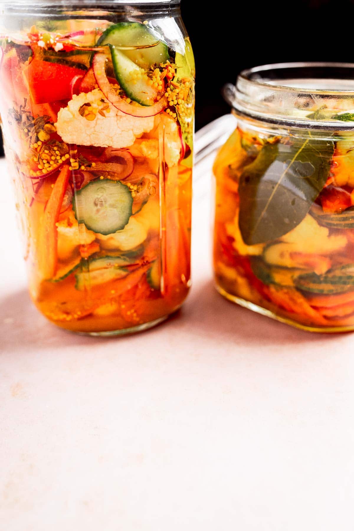 Two glass jars filled with vibrant pickled vegetables, like cucumbers, carrots, and red peppers, reminiscent of torshi, with herbs floating in a brine. The jars grace a light surface.