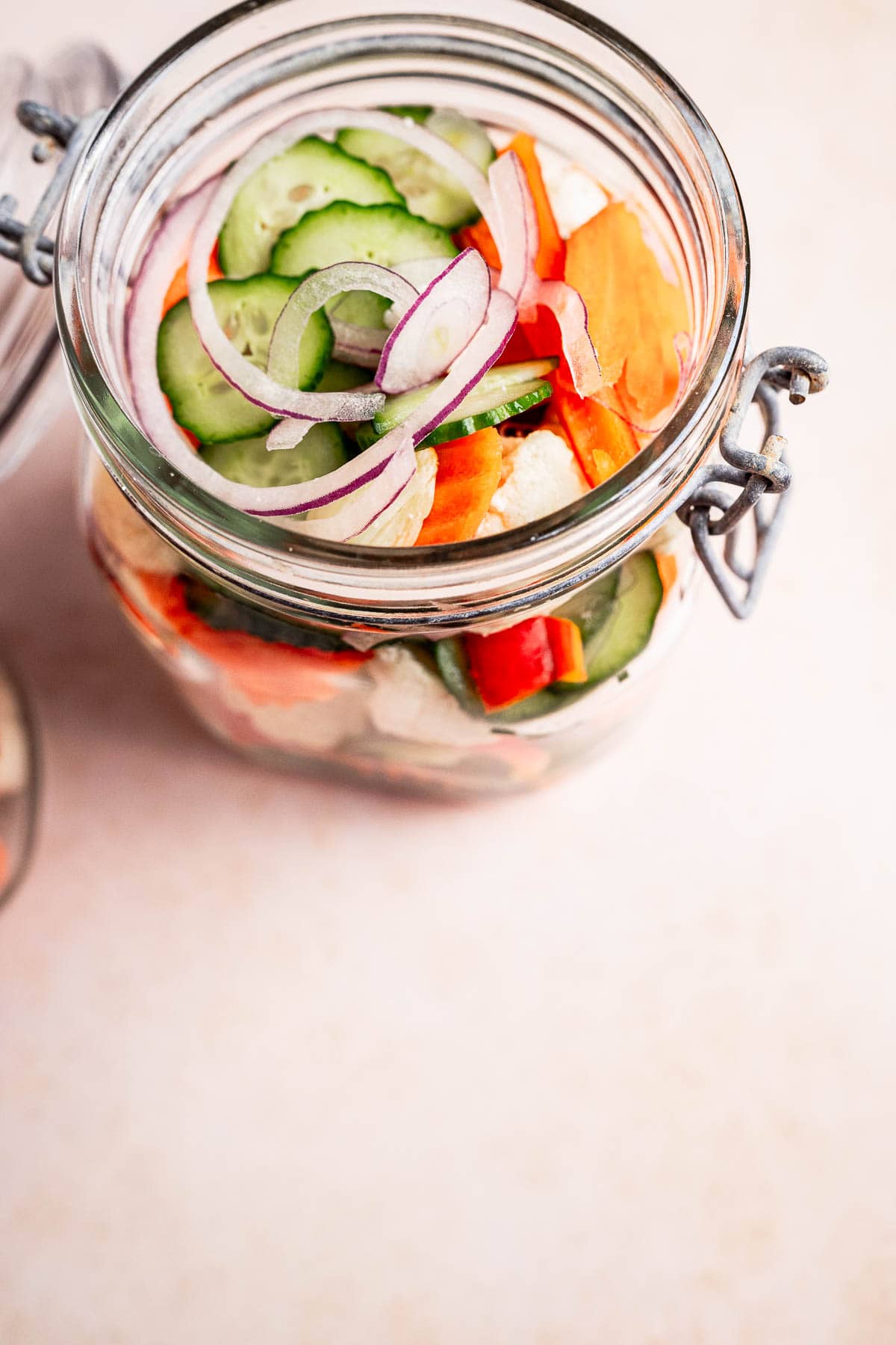 The open glass jar is brimming with vibrant torshi, featuring a colorful mix of sliced cucumbers, onions, carrots, and bell peppers resting elegantly on a light surface.