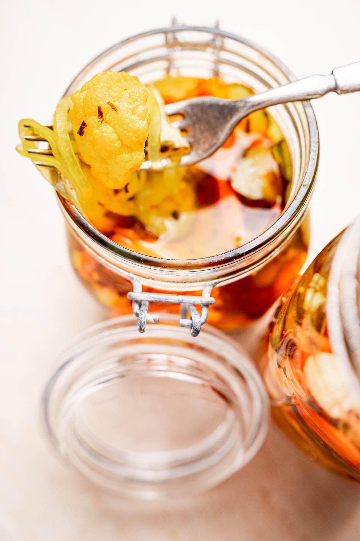 A fork holds vibrant pickled vegetables, or torshi, over an open glass jar brimming with assorted delights like cauliflower and green peppers against a light background.
