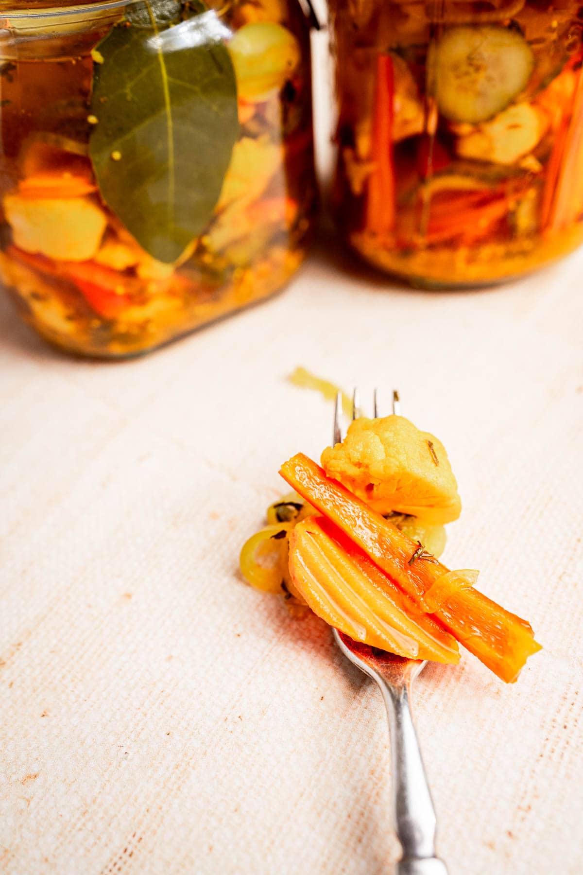 A fork holds tangy pickled vegetables like carrots and cucumbers, with jars of homemade torshi glistening in the background on a light surface.