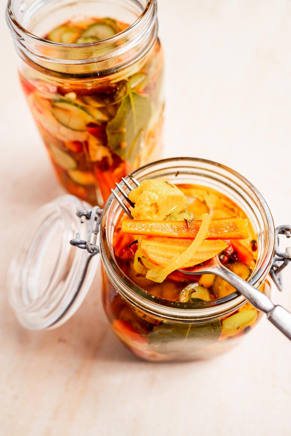 Two jars of torshi, featuring pickled vegetables like carrots and cauliflower. One jar is open, with a fork holding some of the vibrant mix above it.
