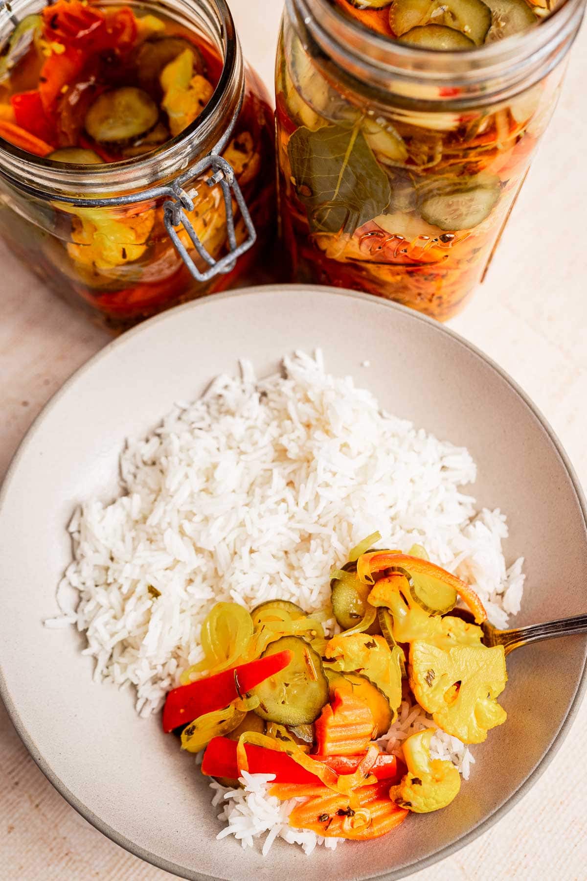 A bowl of white rice topped with torshi, including pickled cucumbers, carrots, and bell peppers. Two jars of similar flavorful pickled vegetables are in the background.