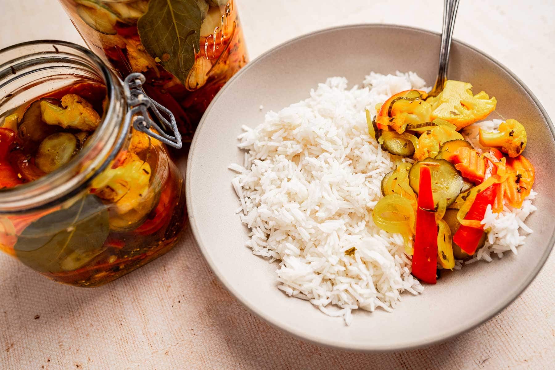 A bowl of white rice with colorful pickled vegetables, or torshi, on the side. Two jars of torshi sit nearby, adding a vibrant touch.