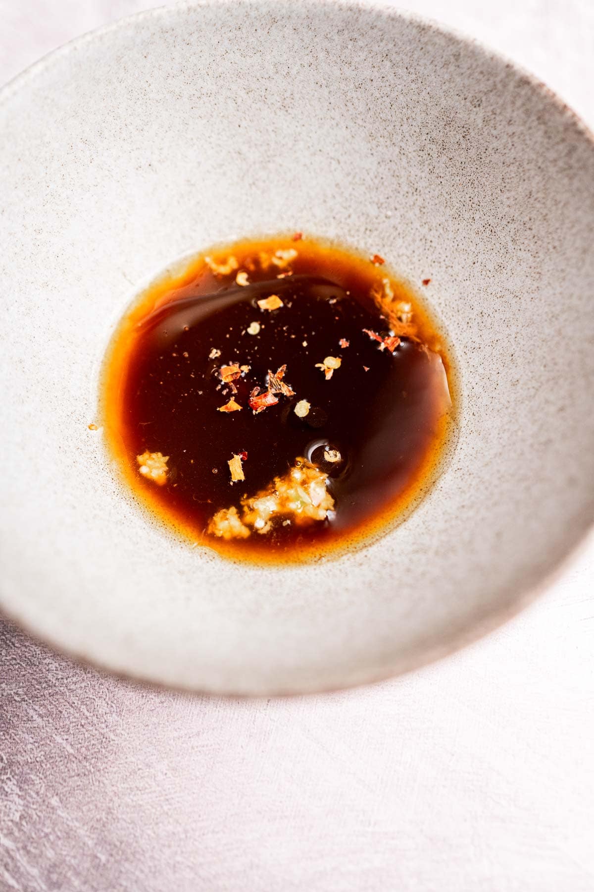A white bowl contains a dark liquid sauce with floating bits of red pepper flakes and minced garlic, perfectly paired with rice paper noodles on a textured surface.