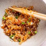 A bowl of jellyfish salad with sesame seeds and chopped green onions, being picked up with chopsticks.