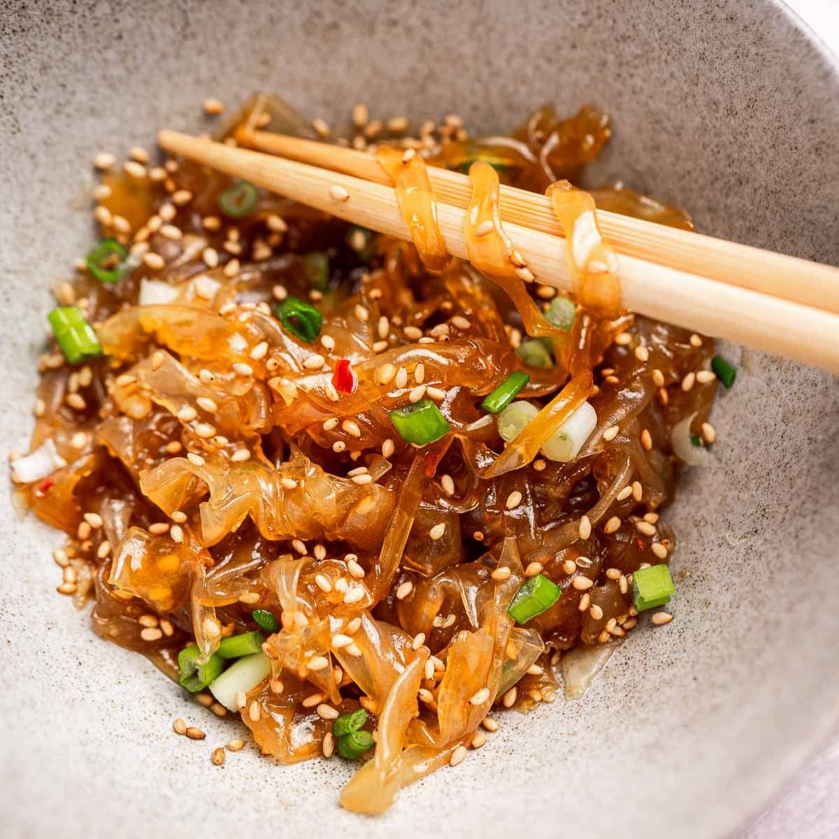 A bowl of jellyfish salad with sesame seeds and chopped green onions, being picked up with chopsticks.