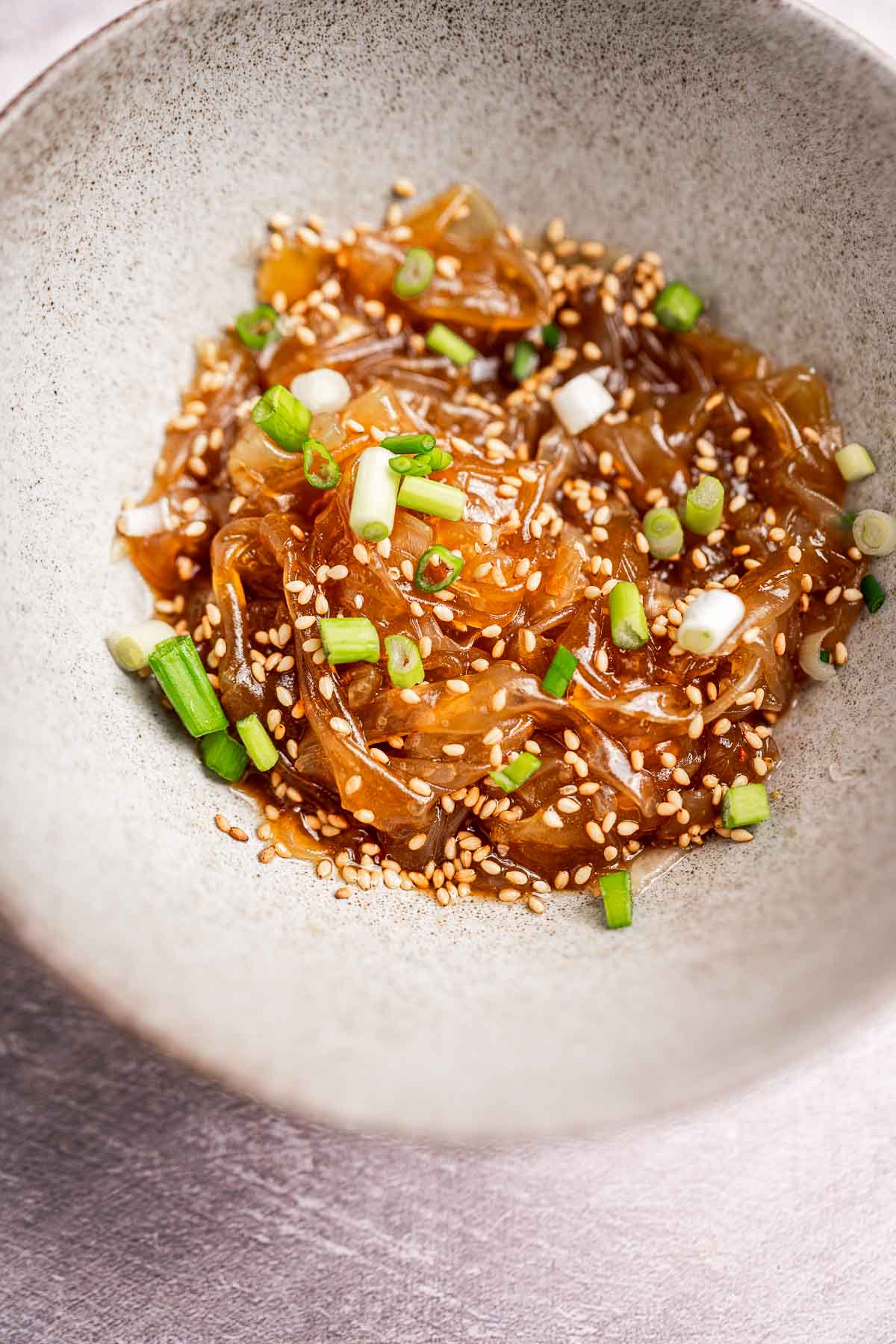 A bowl of marinated jellyfish salad served over rice paper noodles, topped with sesame seeds and chopped green onions.