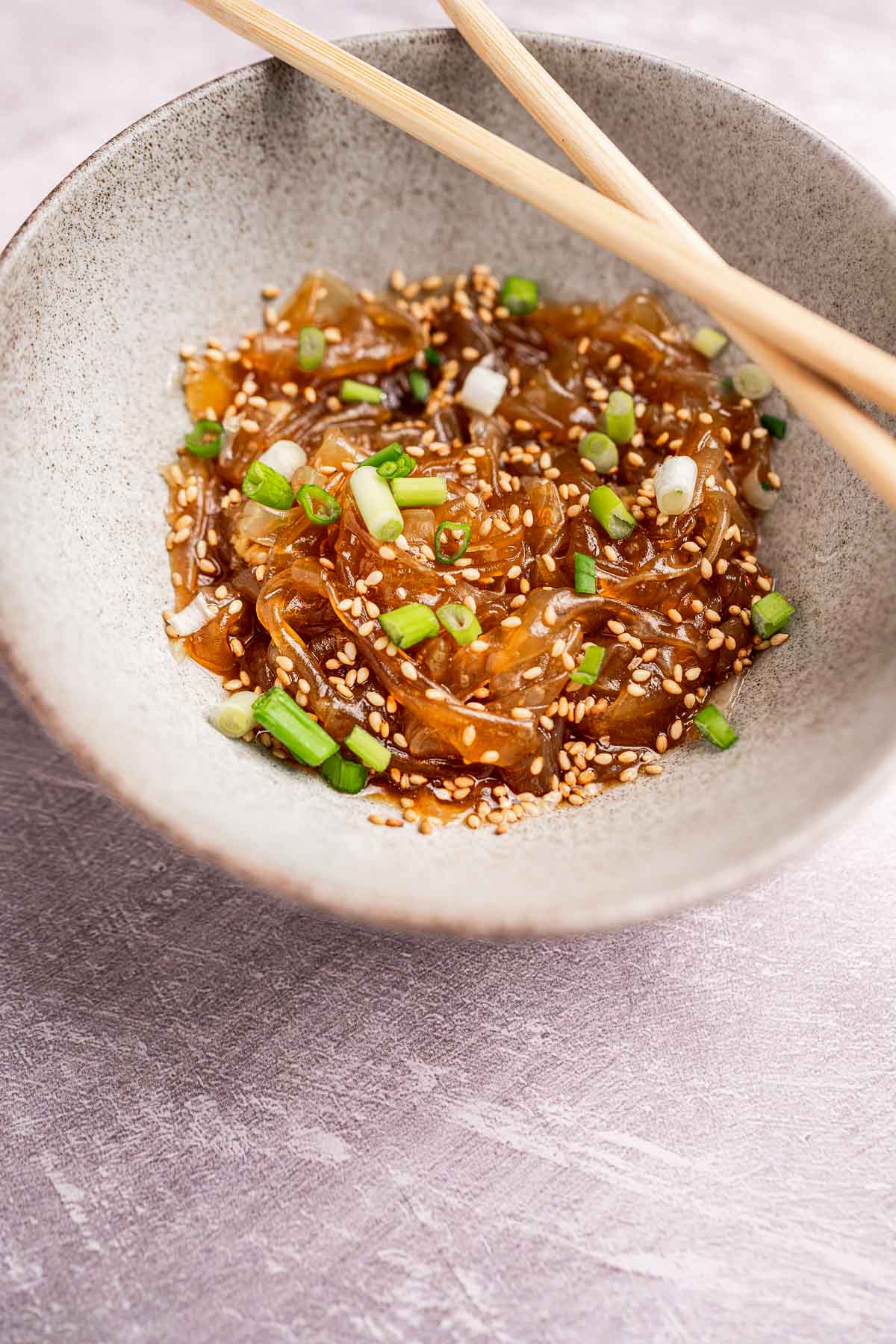 A bowl of seaweed salad garnished with sesame seeds and chopped green onions, accompanied by tender rice paper noodles, with chopsticks resting on the side.