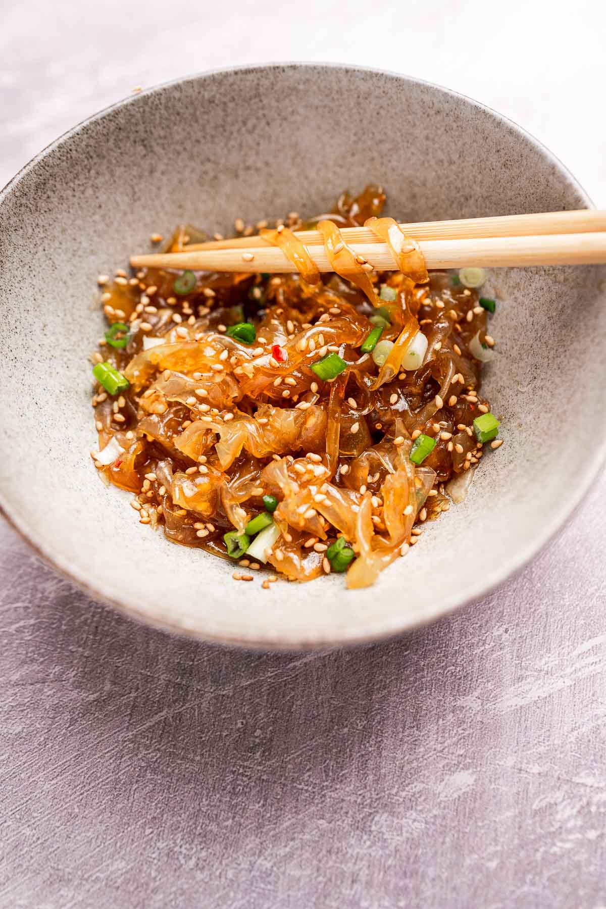 A bowl of stir-fried onions with sesame seeds, chopped green onions, and chili flakes, served alongside delicate rice paper noodles and chopsticks.
