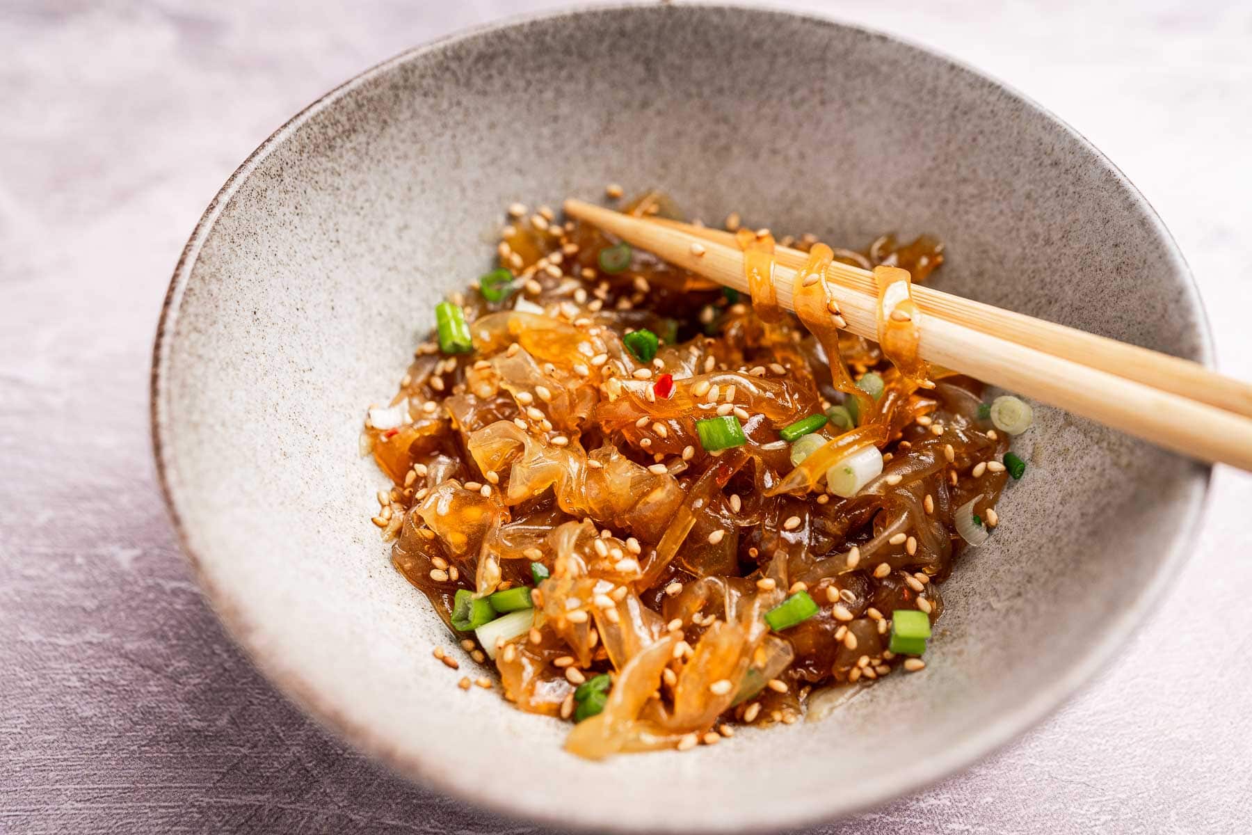 A bowl of jellyfish salad with rice paper noodles, sprinkled with sesame seeds and chopped green onions, with chopsticks resting on the rim.
