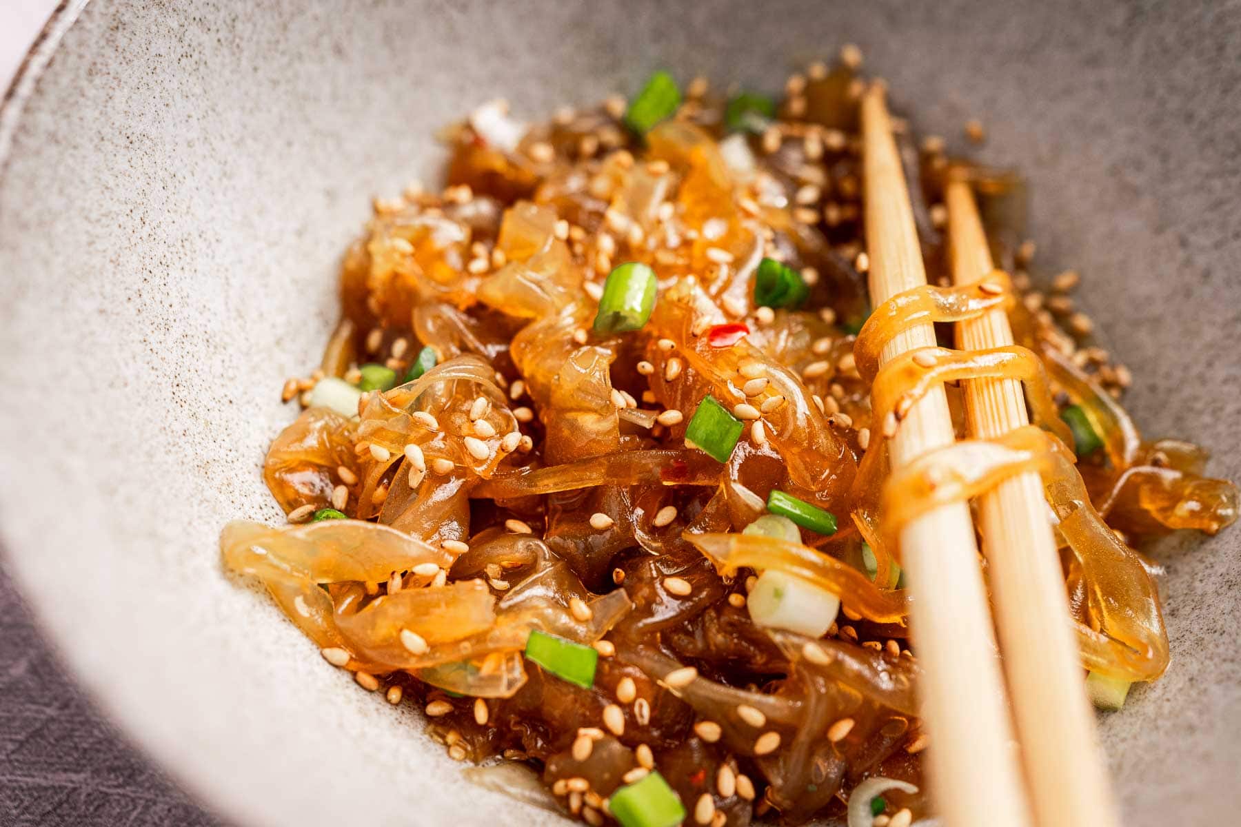 Bowl of jellyfish salad with rice paper noodles, topped with sesame seeds and chopped green onions, accompanied by wooden chopsticks.