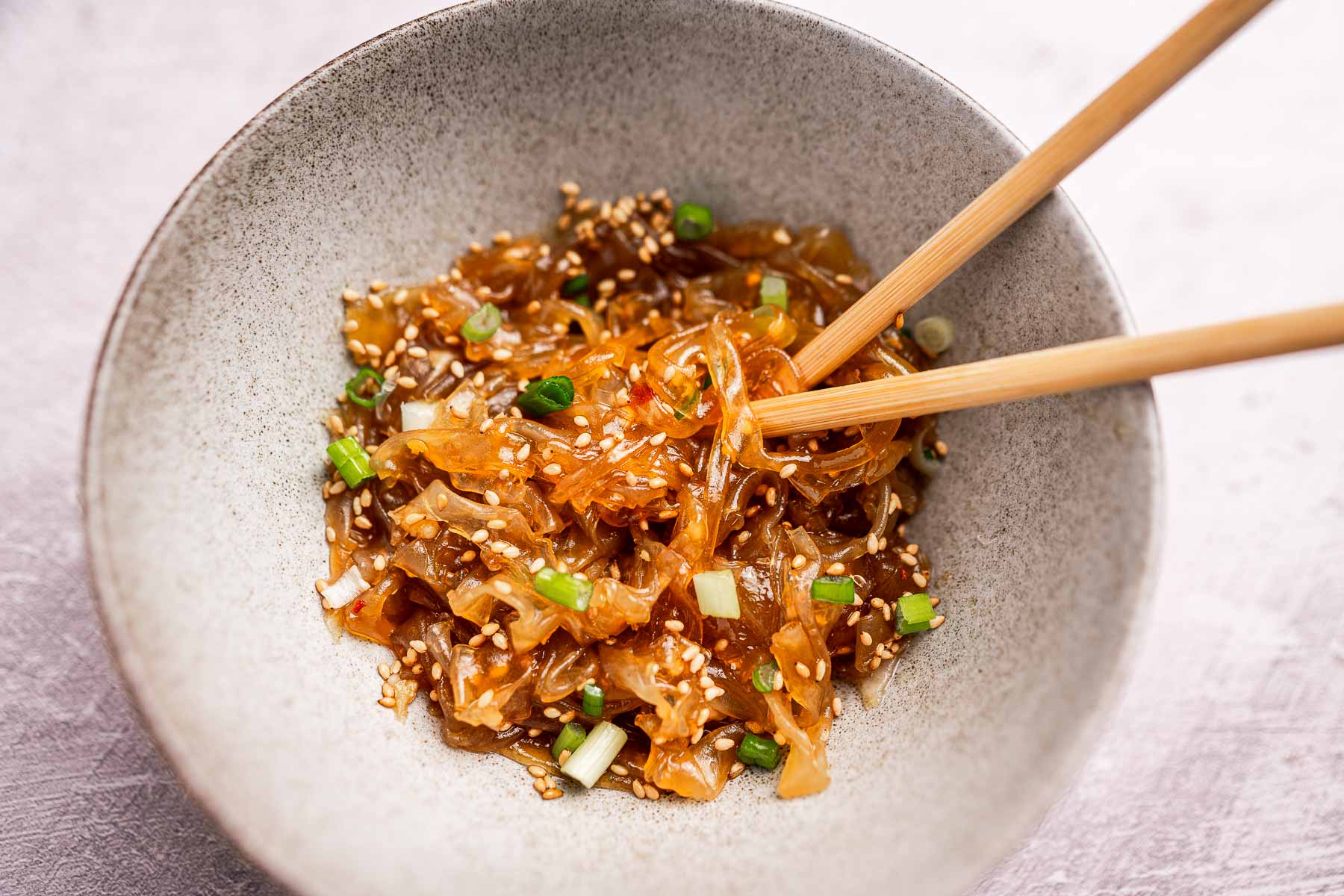 A bowl of seasoned rice paper noodles garnished with sesame seeds and chopped green onions, with chopsticks resting on the edge.