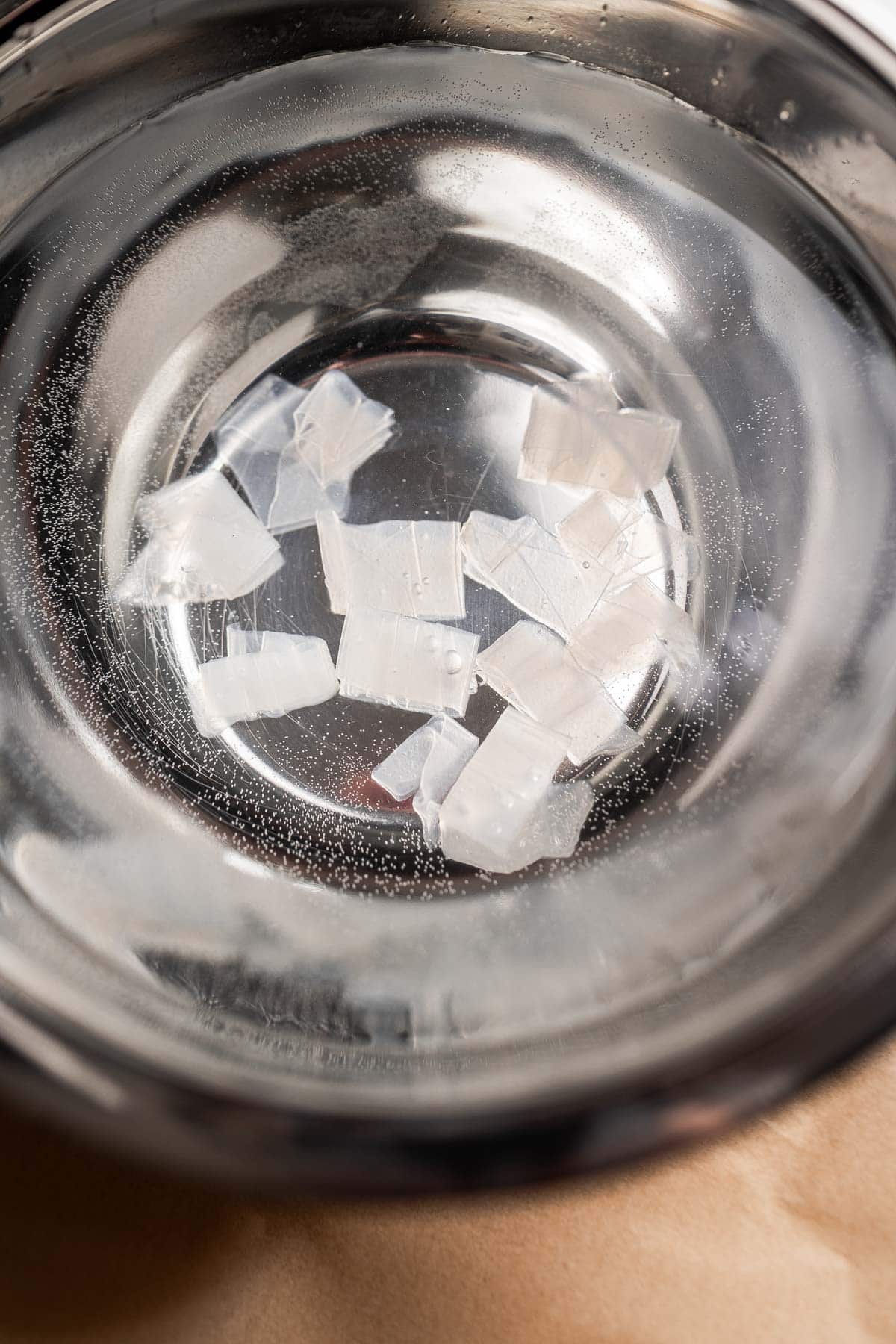 A stainless steel bowl containing water, several small ice cubes, and a scattering of rice paper noodles.