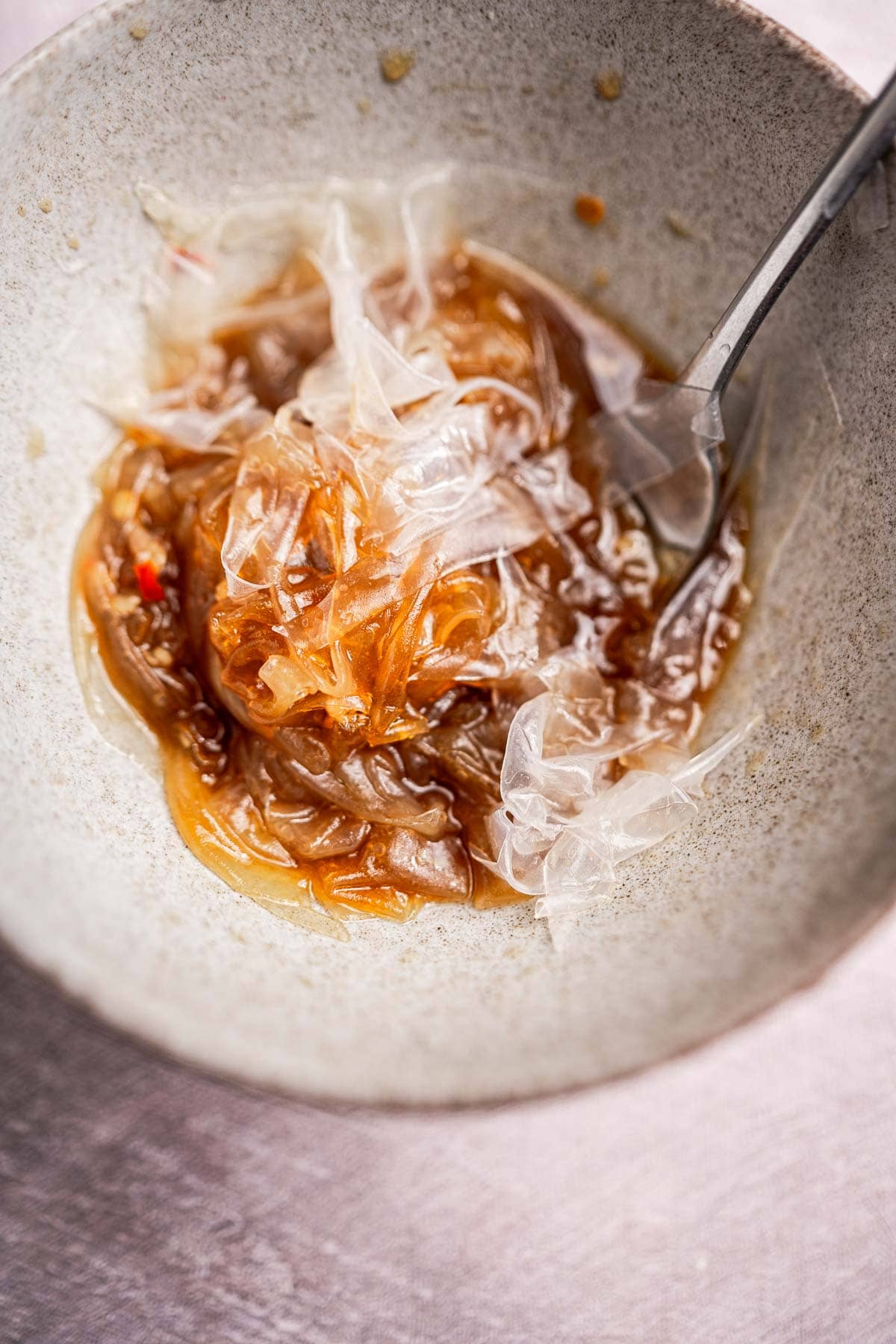 Bowl containing jellyfish salad with a fork, featuring translucent strips in a light brown dressing, complemented by delicate rice paper noodles.
