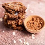Stack of oatmeal cookies with salt flakes beside a wooden bowl of cocoa powder on a wooden surface.