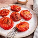 Plate of roasted tomato slices with seasoning, next to a fork. Bowl with salt in the background.