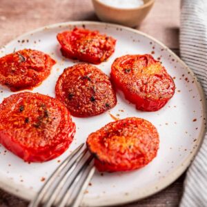 Plate of roasted tomato slices with seasoning, next to a fork. Bowl with salt in the background.