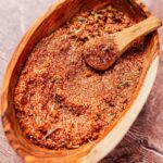 A wooden bowl filled with a reddish-brown spice mix and a small wooden scoop on a wooden surface.