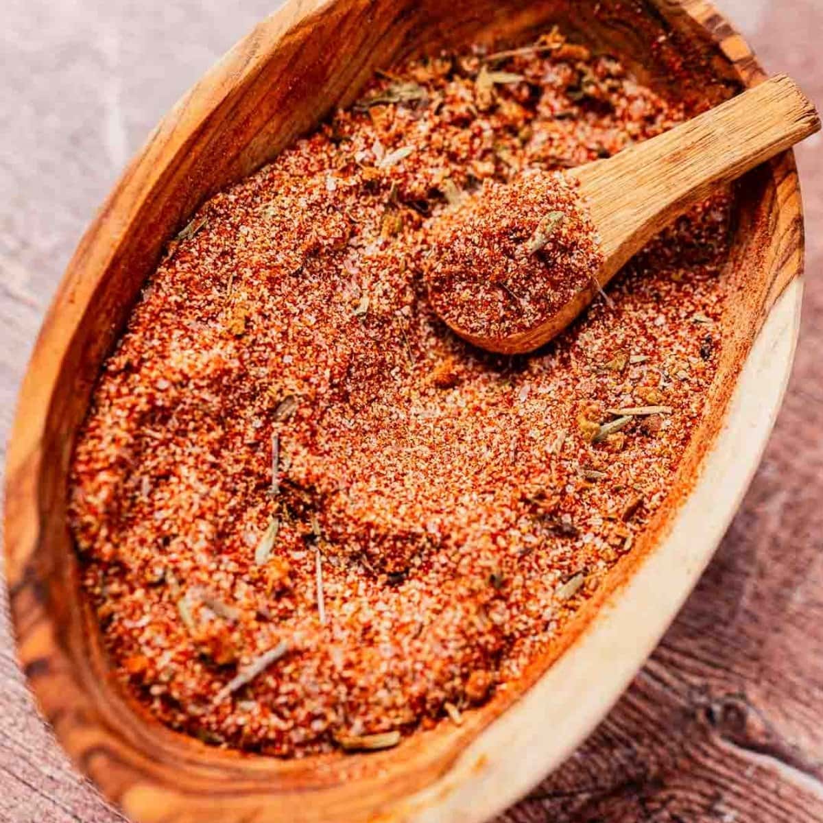 A wooden bowl filled with a reddish-brown spice mix and a small wooden scoop on a wooden surface.