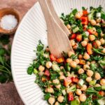 A bowl of chopped chickpea salad brimming with red peppers, cucumbers, and parsley. Two wooden utensils rest inside the vibrant mix. A small bowl of salt waits nearby for a dash of extra flavor.