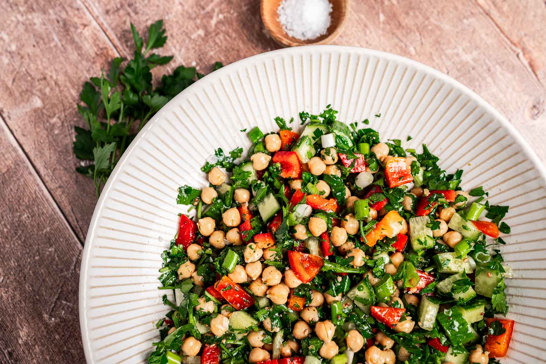 A bowl of chopped chickpea salad brimming with diced red peppers, cucumbers, tomatoes, and parsley sits on a wooden surface. In the background, a small dish of salt awaits to enhance the vibrant flavors.